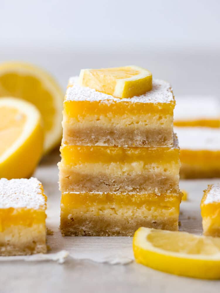 These are the most delicious and creamy lemon cheesecake bars. They have a buttery shortbread crust with a cheesecake and lemon layer and are dusted with powdered sugar. They are the perfect dessert! A stack of lemon cheesecake bars with a lemon slice on top.