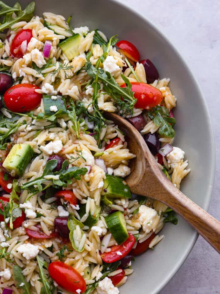 If you are looking for a delicious salad full of flavor and packed with different textures, then this Greek orzo salad is for you! Vegetables and arugula drizzled with a lemon vinaigrette combine to make the most delicious and refreshing taste! A close up of a bowl of Greek orzo salad with a wooden spoon.
