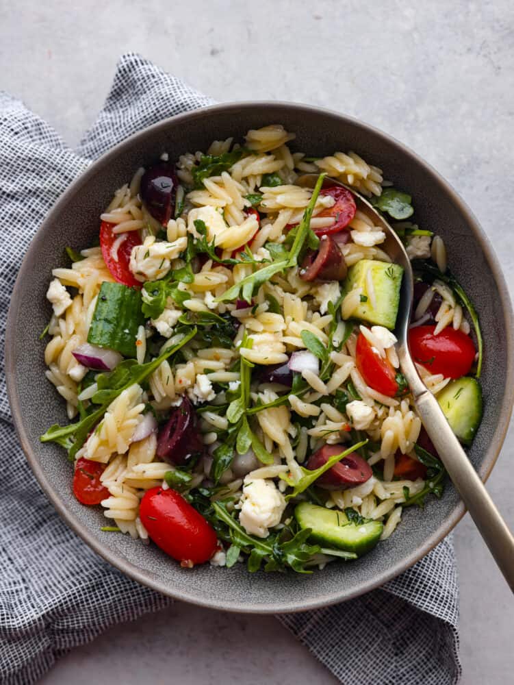 If you are looking for a delicious salad full of flavor and packed with different textures, then this Greek orzo salad is for you! Vegetables and arugula drizzled with a lemon vinaigrette combine to make the most delicious and refreshing taste! The top view of a serving size bowl filled with salad and a spoon.