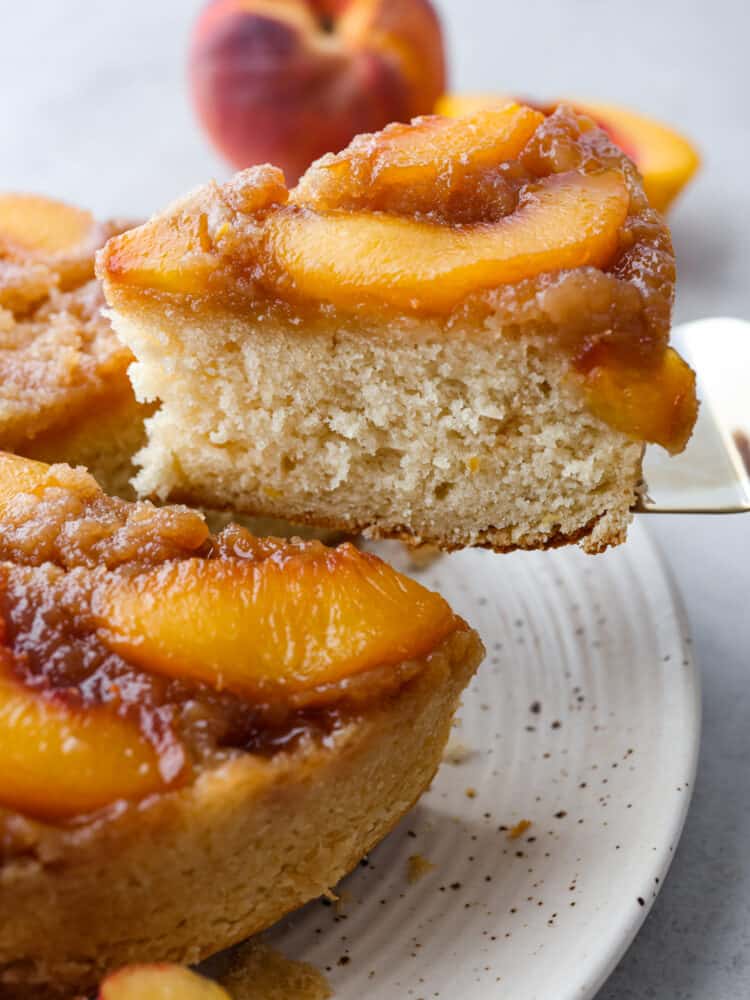 Get ready for your mouth to water as I tell you about this amazingly delicious peach upside-down cake! It is juicy, full of sweet flavor from brown sugar crumbles, and the best part... The soft, melt-in-your-mouth cake that combines it all together! The side view of a slice of peach upside down cake being taken out with a cake server.