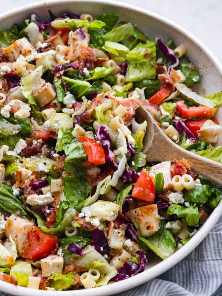 This copycat portillo's chopped salad is the best combination of all time! Romaine lettuce seasoned with a delicious and flavorful vinaigrette dressing. Topped with some pasta, veggies, and cheese, you can't forget the best part... the bacon! A bowl of Portillo's chopped salad with a wooden spoon in it, ready to serve it.