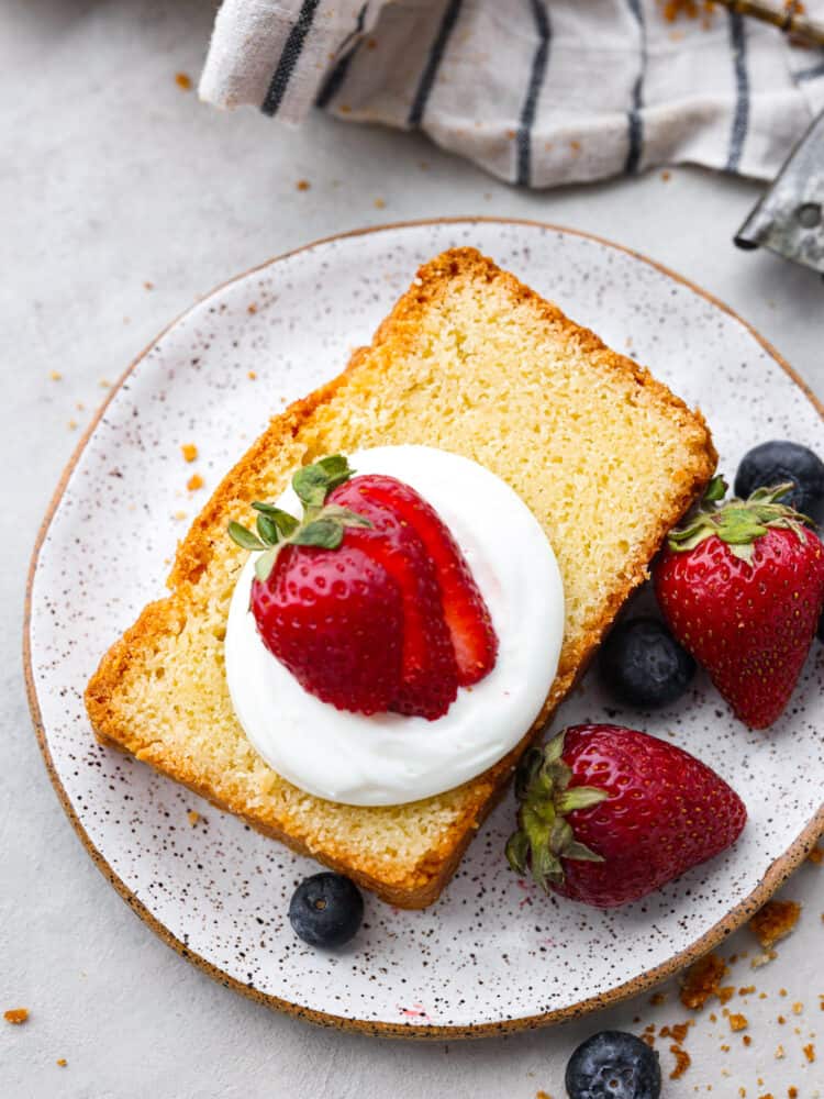 This is a classic recipe that really IS the perfect pound cake! There is something about this exact mix of these simple pantry staple ingredients that results in a velvety smooth and perfectly dense and tender cake. A slice of cake on a stone plate with whipped cream and strawberries.