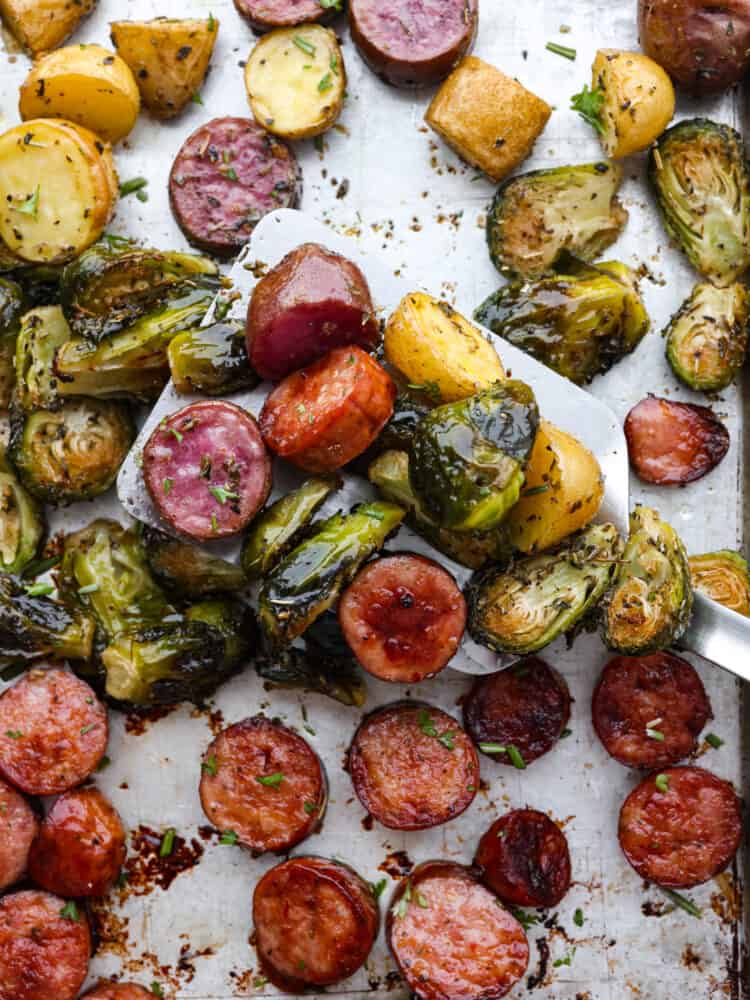 This sheet pan sausage and veggies are straight from heaven! This recipe has potatoes, sausage, and Brussel sprouts that are all perfectly seasoned with dried Italian herbs. You will want to eat every last bite! A close up of sausage and veggies on a sheet pan being scooped up with a spatula.