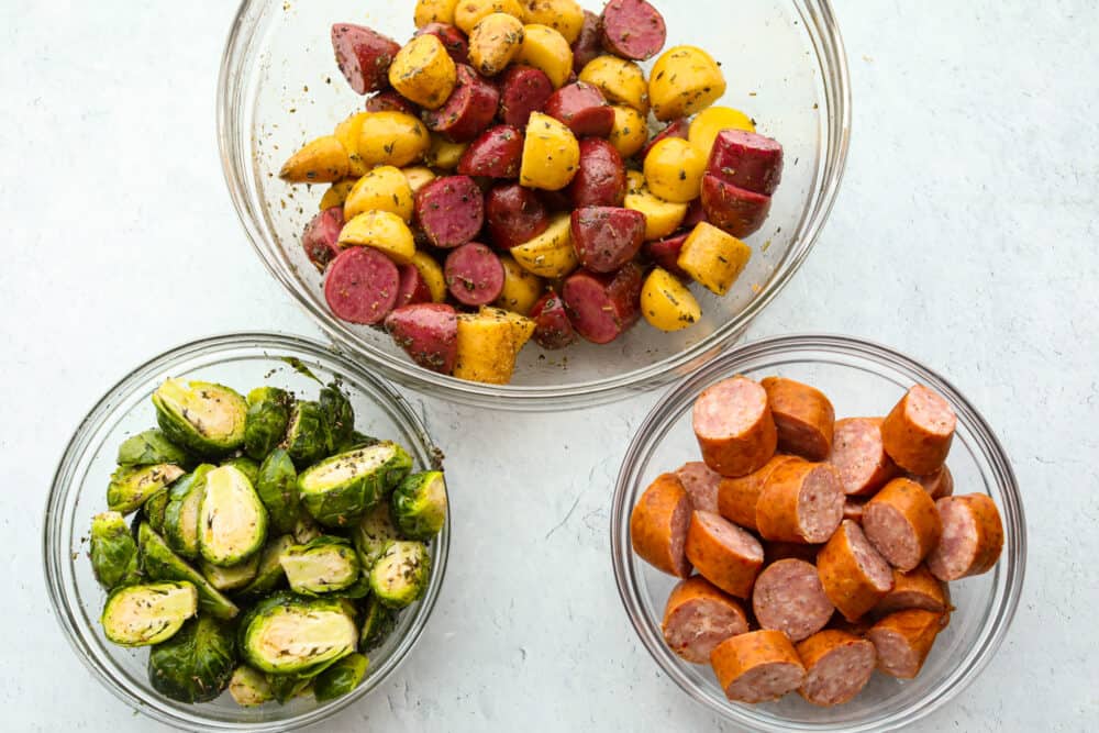 This sheet pan sausage and veggies are straight from heaven! This recipe has potatoes, sausage, and Brussel sprouts that are all perfectly seasoned with dried Italian herbs. You will want to eat every last bite! A picture showing 3 glass bowls. One has cut up potatoes, one has sliced Brussel sprouts and the other has sliced sausage.
