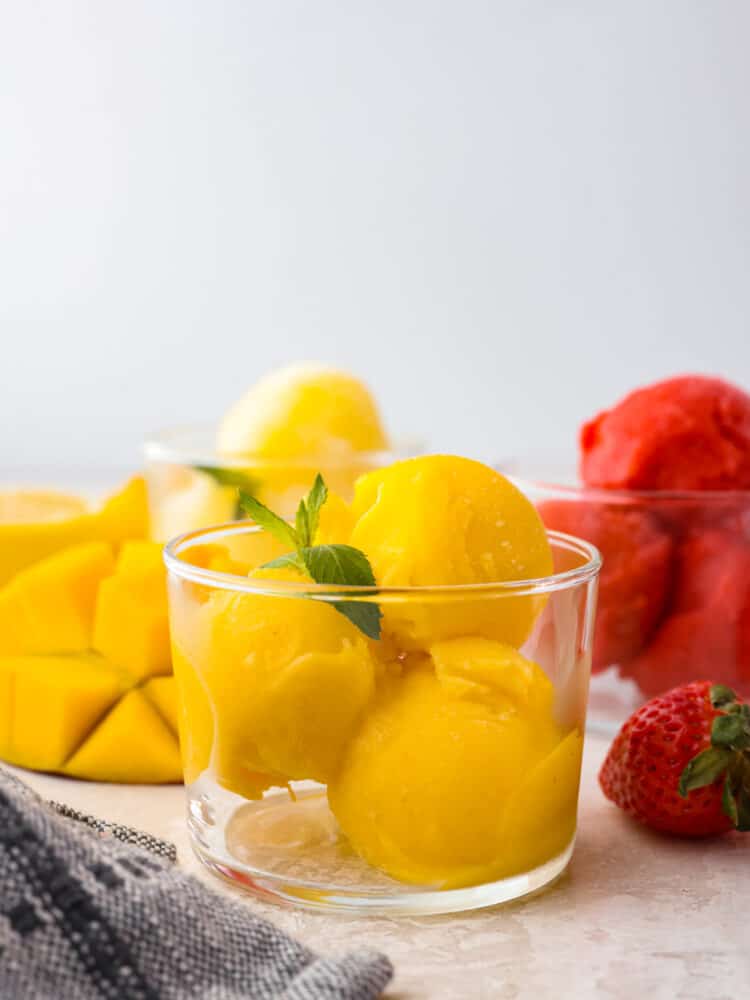 Get ready for the most refreshing homemade sorbet that you have ever had with this perfect recipe! Silky smooth, packed full of flavor, and a delightful dessert for any occasion, this will knock your socks off! A glass cup with a few scoops of homemade mango sorbet with a mint leaf on top.