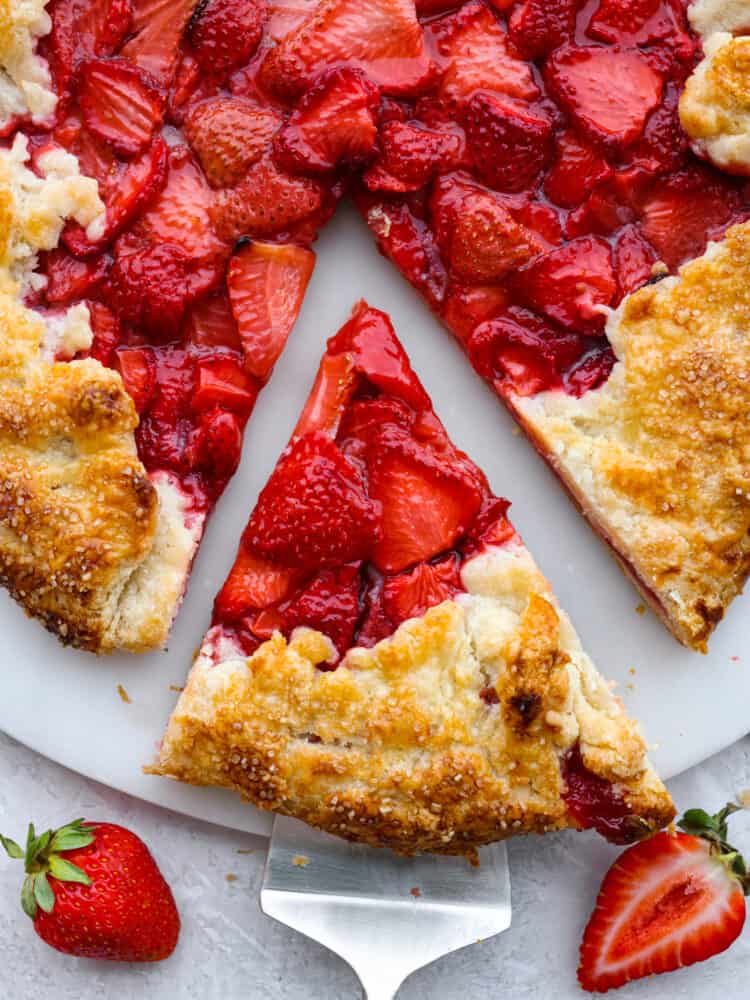 This fresh strawberry galette uses sweetened strawberries that are wrapped in a beautiful rustic buttery pastry and baked to perfection. Strawberry galette is a much easier version of a pie, and it will become your new favorite strawberry dessert! Close up and top view of a slice of galette being served. Fresh strawberries garnished on the side.