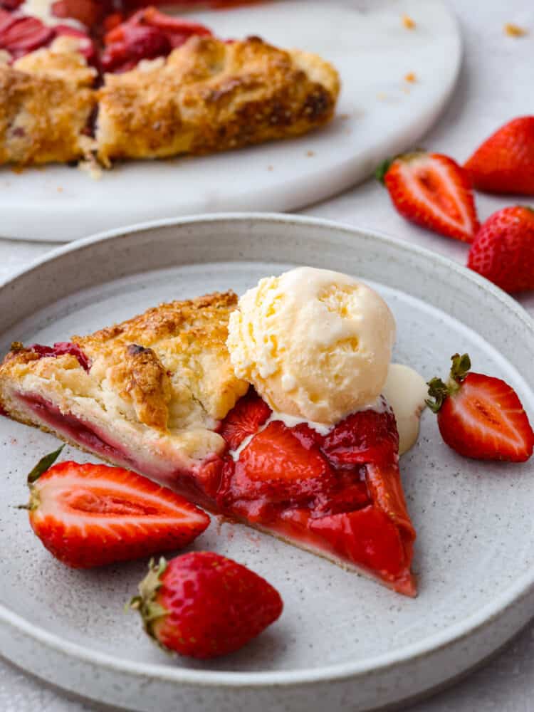 Fresh sweetened strawberries wrapped in a beautiful rustic buttery pastry and baked to perfection. Strawberry galette is a much easier version of a pie, and it will become your new favorite strawberry dessert! A slice of strawberry galette on a gray plate topped with a scoop of ice cream, and garnished with fresh strawberries on the side. A view of the remaining galette is pictured in the distance.
