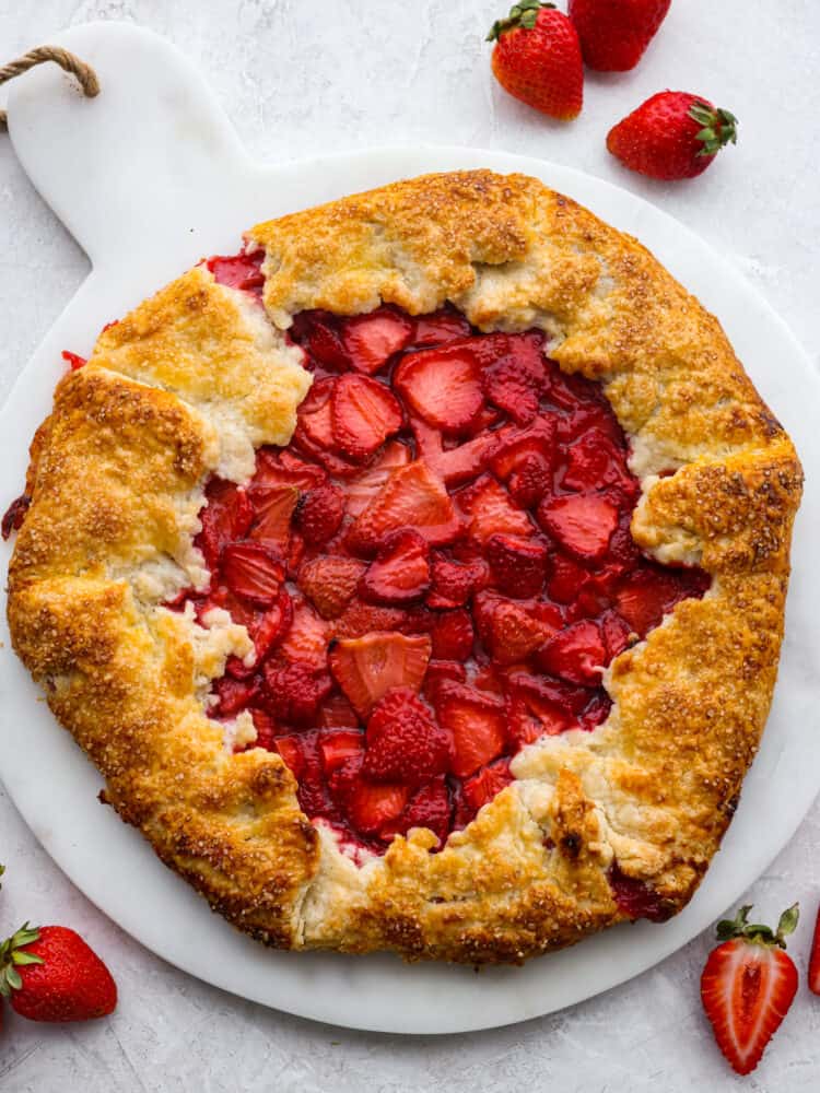 This fresh strawberry galette uses sweetened strawberries that are wrapped in a beautiful rustic buttery pastry and baked to perfection. Strawberry galette is a much easier version of a pie, and it will become your new favorite strawberry dessert! Top view of baked strawberry galette on a marble board. Fresh whole and halved strawberries scattered beside the galette.