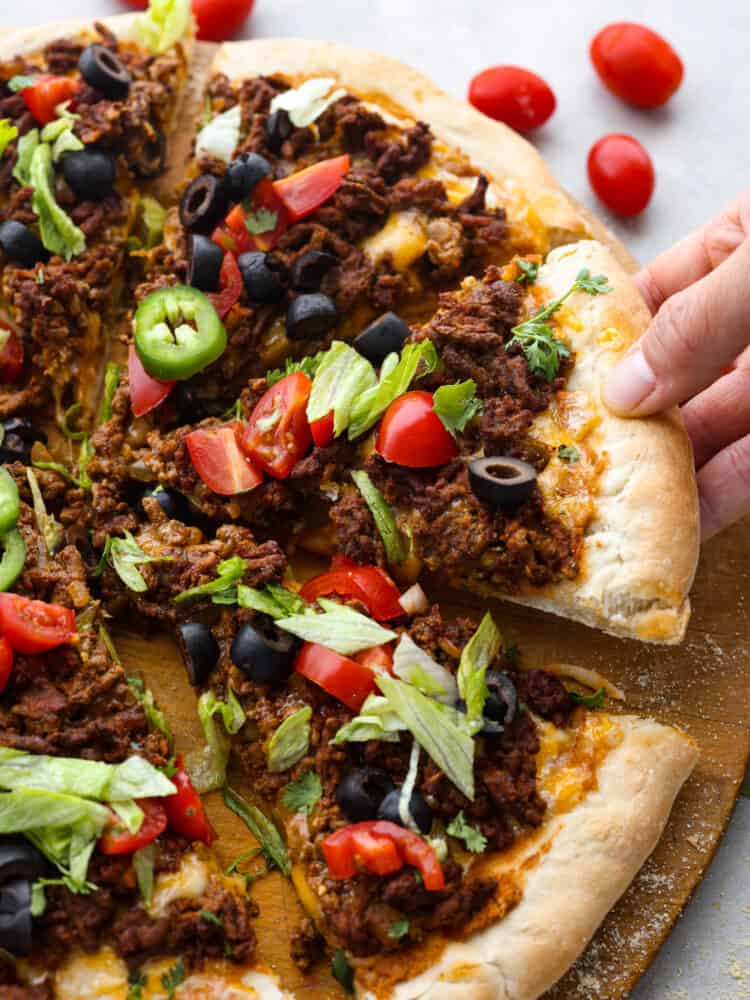 Two classic family favorites, pizza and tacos, come together in this easy weeknight meal that is destined to become a favorite in your home. Taco pizza is loaded with tasty seasoned beef, melty cheese, beans, and fresh veggies for a flavor combination you'll love! A slice of taco pizza being picked up off of the serving board.