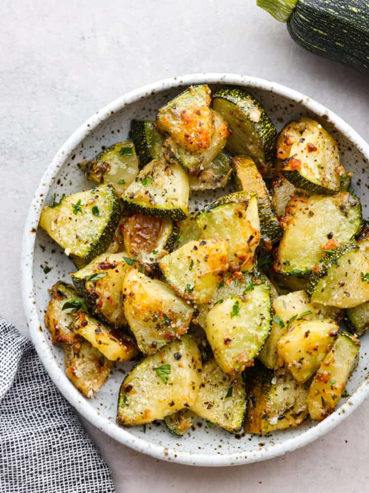 If you have never cooked air fryer zucchini, then you are in for the tastiest treat! It's perfectly seasoned with Italian seasonings and parmesan cheese and cooked to perfection! A whte speckled bowl filled with cooked zucchini slices.