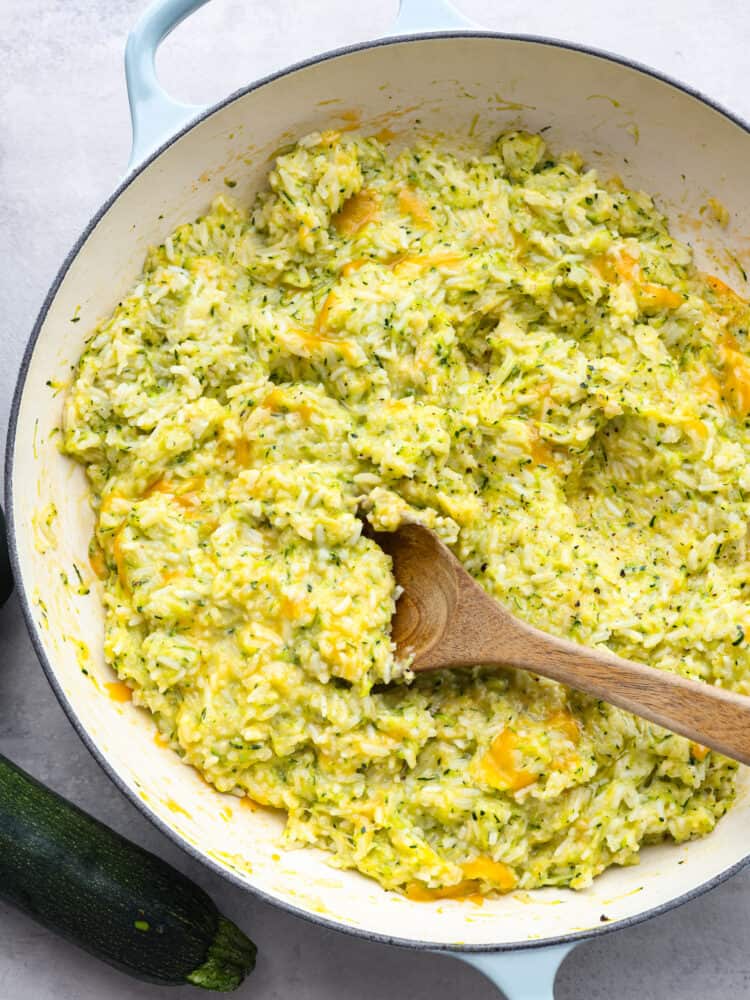 This cheesy zucchini rice is ooey gooey and a divine comfort food that makes the best side to any meal. It is so cheesy and delicious, and the best part is that it has a vegetable in it that my kids will actually eat! The top view of a pan filled with cooked cheesy zucchini rice and a wooden spoon.