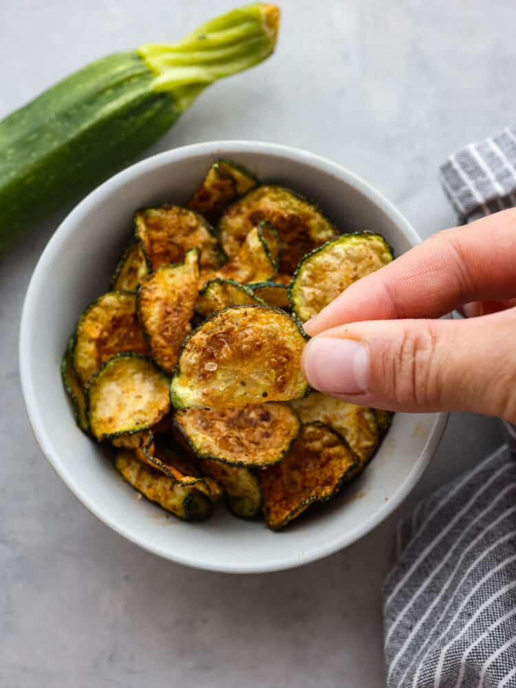 If you're looking for a healthy and delicious way to use up your summer zucchini, making chips in the air fryer is the answer! They're low-carb, seasoned to perfection, and perfectly golden and crispy! A zucchini chip being picked up out of a small white bowl.