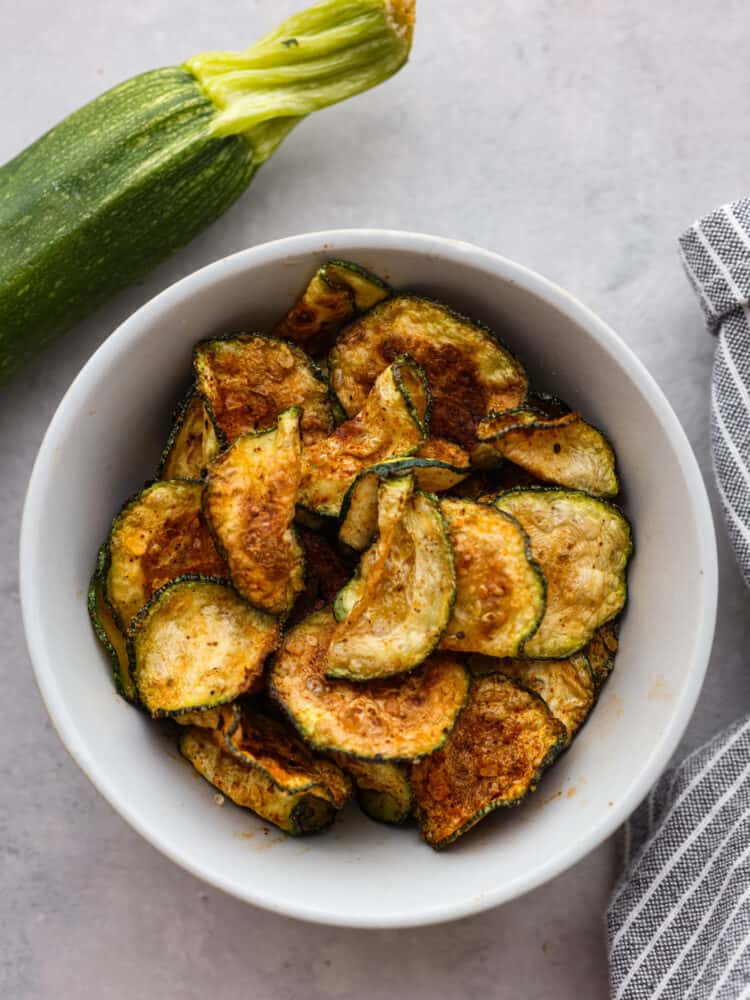 If you're looking for a healthy and delicious way to use up your summer zucchini, making chips in the air fryer is the answer! They're low-carb, seasoned to perfection, and perfectly golden and crispy! Top-down view of air-fried zucchini chips in a white bowl.