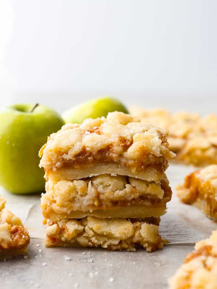 Salted caramel apple bars are ooey-gooey treats that have an amazing buttery shortbread crust and a soft and sweet caramel apple center!ย This is one of the best desserts you will make this fall! A stack of caramel apple bars on the counter with some green apples in the background.