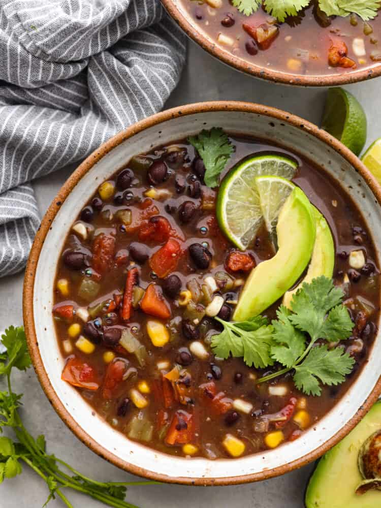 A super easy 20-minute black bean soup! This soup is hearty and filling and a great option for those wanting a vegetarian option! It's made with black beans, a variety of veggies, and delicious seasonings for a savory soup with a hint of warm spice! Top-down view of black bean soup in a speckled stoneware bowl, garnished with lime, avocado slices, and fresh cilantro.