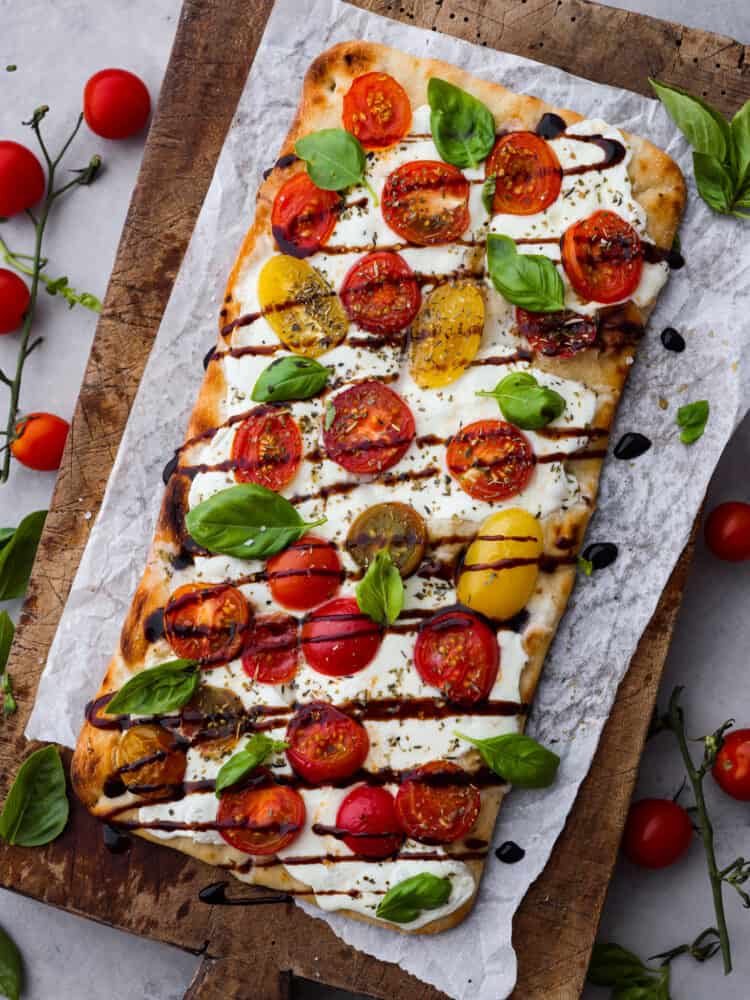 Light and fresh, this caprese flatbread is the perfect appetizer for any Italian-inspired meal! It’s topped with melty mozzarella, fresh herbs and veggies, and topped off with a mouthwatering balsamic glaze! A full caprese flatbread on parchment paper.