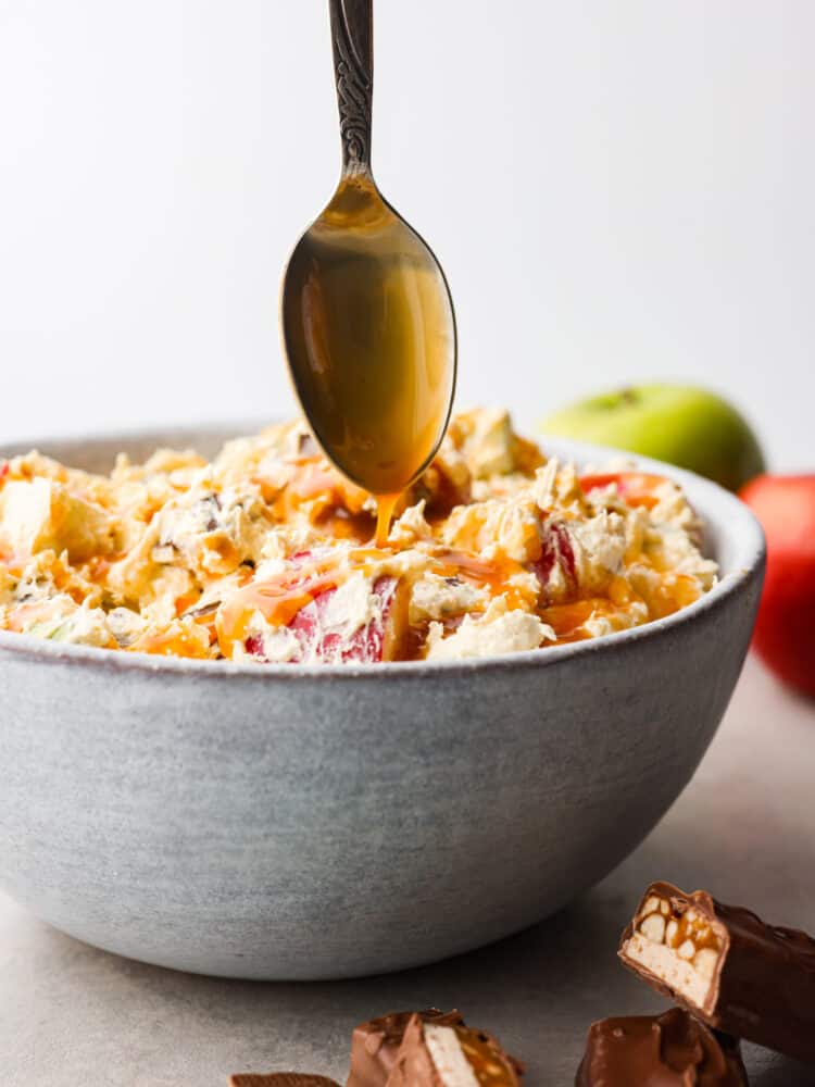 Snickers salad is perfect for any potluck! It's a mixture of snickers bites and apples, all covered in a vanilla cool whip and topped with chocolate and caramel sauce. This perfectly sweet and salty salad is so delicious and surprisingly easy to make! Snickers salad in a bowl with a spoon drizzling on some caramel sauce.