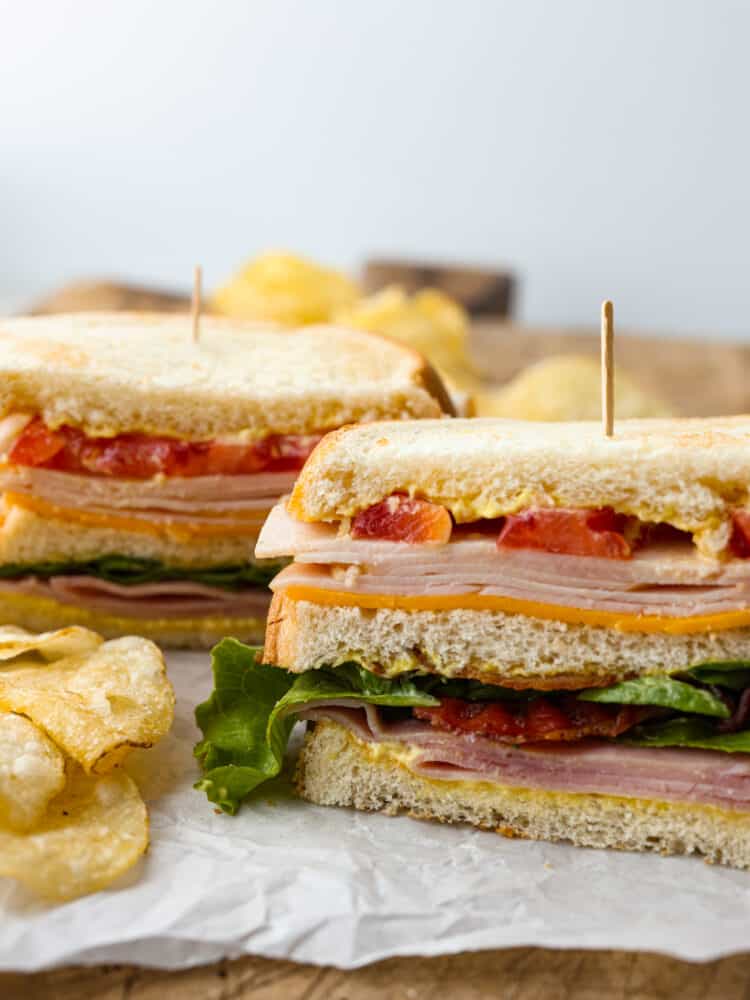 There’s a reason why club sandwiches are so popular - you can’t go wrong with 3 pieces of toasty bread smothered in mayonnaise, mustard, and then layered with meat, cheese, and fresh greens! This classic club sandwich recipe is going to be your new lunch go-to. Front view of 2 club sandwiches filled with meat, cheese, and vegetables, held together by toothpicks.