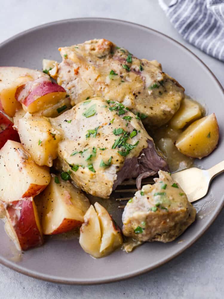 Creamy ranch pork chops are the perfect go-to weeknight meal. Tender pork chops and red potatoes are covered in a creamy, zesty sauce in this 5-ingredient crock pot recipe! Closeup of ranch pork chops and potatoes served on a gray plate. The pork chop has a slice cut out of it.