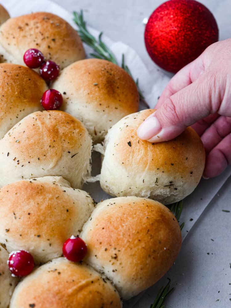 Easy, delicious, and adorably festive, this Dinner Roll Wreath is perfect for holiday dinners! Warm and soft rolls arranged to look like a wreath are sure to spruce up your holiday dinner table. A close up of dinner roll wreath with a hand grabbing one of the rolls.