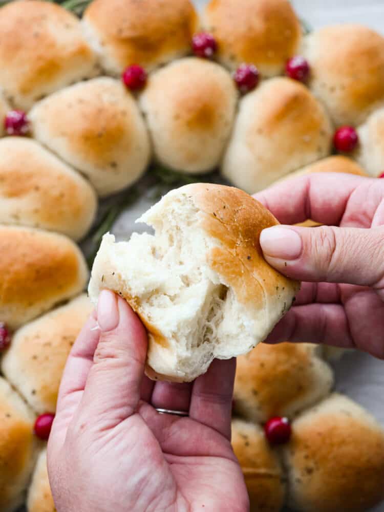 Easy, delicious, and adorably festive, this Dinner Roll Wreath is perfect for holiday dinners! Warm and soft rolls arranged to look like a wreath are sure to spruce up your holiday dinner table. Two hands ripping open a dinner roll with the full dinner roll wreath in the background.