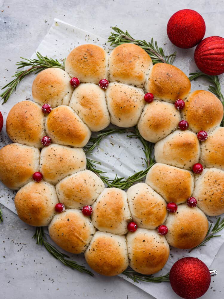 Easy, delicious, and adorably festive, this Dinner Roll Wreath is perfect for holiday dinners! Warm and soft rolls arranged to look like a wreath are sure to spruce up your holiday dinner table. Dinner rolls arranged like a wreath with cranberries on top and rosemary sprigs to the side and red sparkly ornaments.