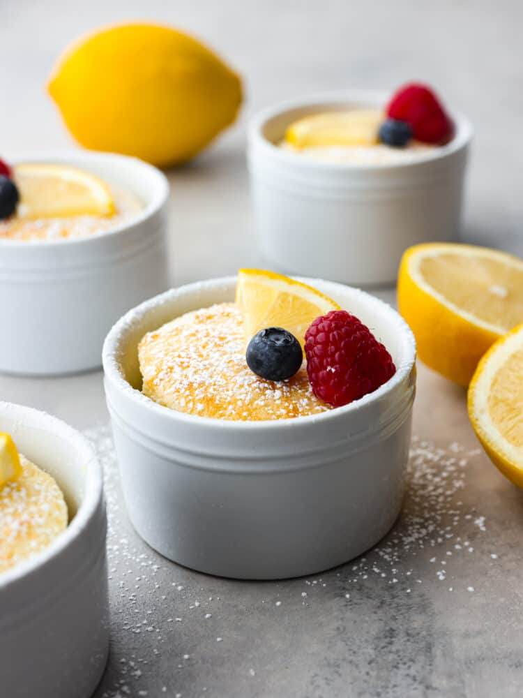 This lemon pudding cake is gorgeous, decadent, and delicious! Dusted with powdered sugar and topped with fresh berries, it's perfect for impressing your guests on any occasion! Lemon pudding cake in individual ramekins with a lemon slice, raspberry and blueberry on top.