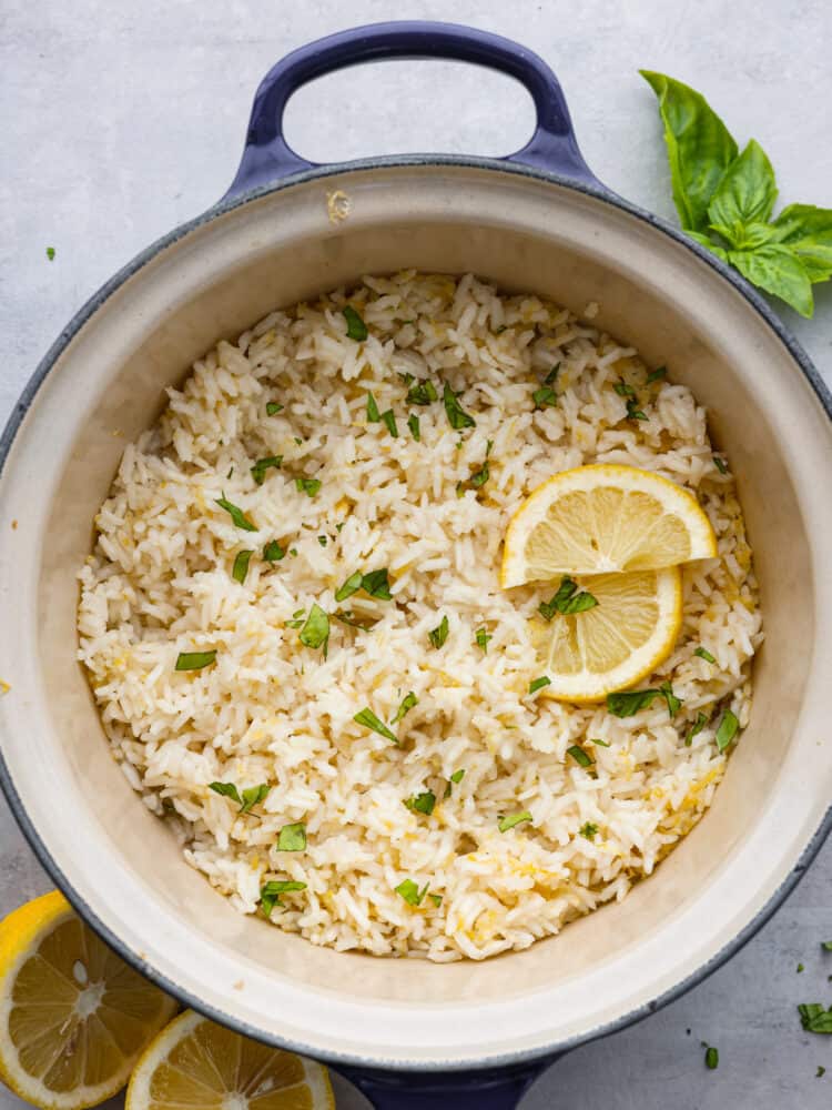 This zesty lemon rice is the perfect summer side dish! It's creamy, savory, and packed with bright citrus flavor! Serve it with your favorite fish or meat dishes for a crave-worthy meal. Top-down view of cooked lemon rice in a blue pot, garnished with chopped basil and lemon slices.