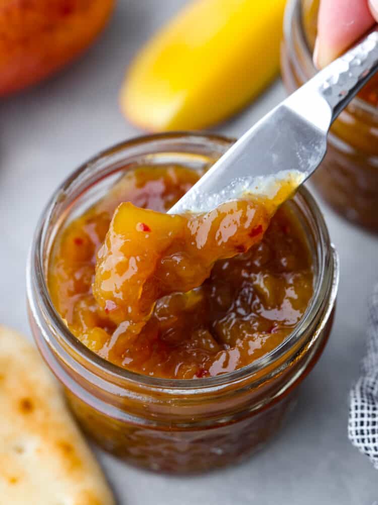 This delightful mango chutney is a spread that can be used in so many different recipes! It packs a punch when it comes to flavor, and you will always want to have some on hand! A jar of mango chutney with a knife getting some out.