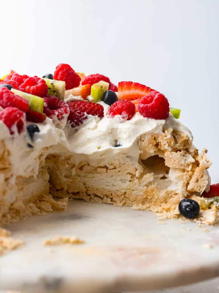 This pavlova has a light and crispy outside with a deliciously soft and chewy inside. Topped with a sweet cream topping and fresh fruit, it's a dessert that will stand out at any gathering! The side view of pavlova with a slice cut out.
