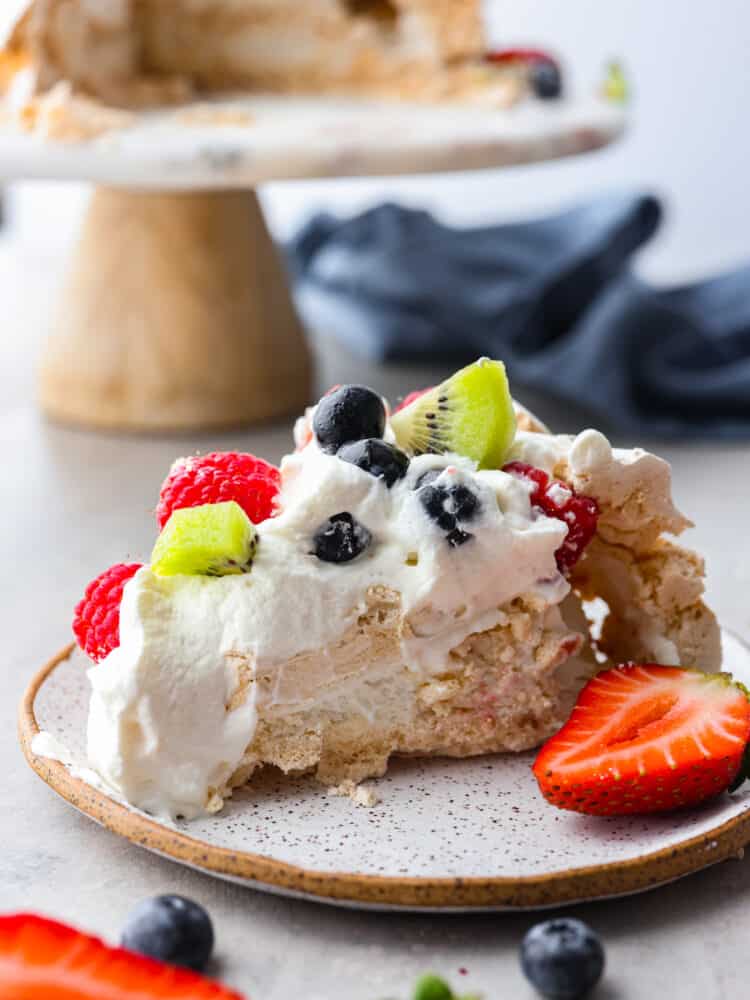 This pavlova has a light and crispy outside with a deliciously soft and chewy inside. Topped with a sweet cream topping and fresh fruit, it's a dessert that will stand out at any gathering! A slice of pavlova on a speckled white plate.