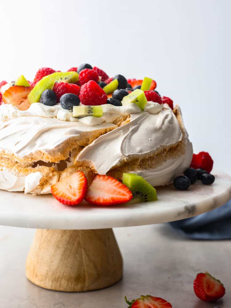 This pavlova has a light and crispy outside with a deliciously soft and chewy inside. Topped with a sweet cream topping and fresh fruit, it's a dessert that will stand out at any gathering! Pavlova on a cake stand topped with fresh fruit.