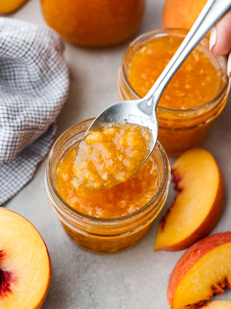 Fresh peach jam can easily be made at home to enjoy all year round. Ripe Summer peaches, sugar, lemon, and pectin quickly turn into a delicious jam with no canning required. Two small glass jars of peach jam surrounded by a kitchen towel, and sliced peaches. A spoon is lifting out a spoonful of jam.