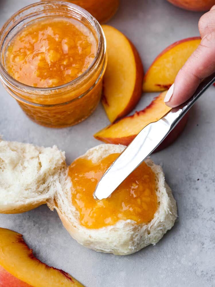 Fresh peach jam can easily be made at home to enjoy all year round. Ripe Summer peaches, sugar, lemon, and pectin quickly turn into a delicious jam with no canning required. A small glass jar of jam surrounded by sliced peaches an an open faced roll. A small knife is spreading jam on the roll.
