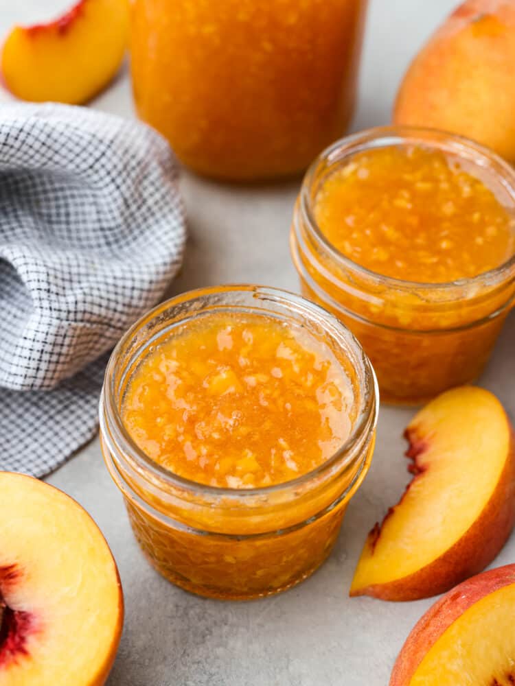 Fresh peach jam can easily be made at home to enjoy all year round. Ripe Summer peaches, sugar, lemon, and pectin quickly turn into a delicious jam with no canning required. Two small glass jars of peach jam surrounded by a kitchen towel, sliced peaches, and a large glass jar of jam.