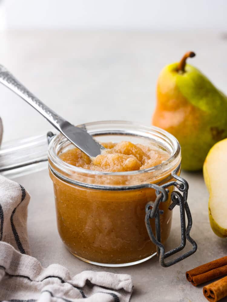 Sweetened with honey and warm spice, this homemade pear butter is a fall staple you'll want to put on everything! It's great for canning and storing to enjoy with all of your favorite baked goods year-round. A glass jar filled with pear butter- cinnamon sticks and whole pears can be seen in the background.