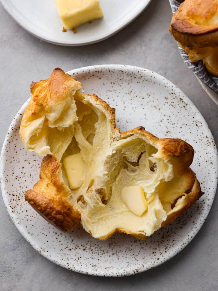 Golden, crisp, and deliciously airy, these homemade popovers are the best addition to any meal! Made with a simple egg batter, popovers have a buttery texture and the perfect chew! A popover torn in half with butter added on a white speckled plate.