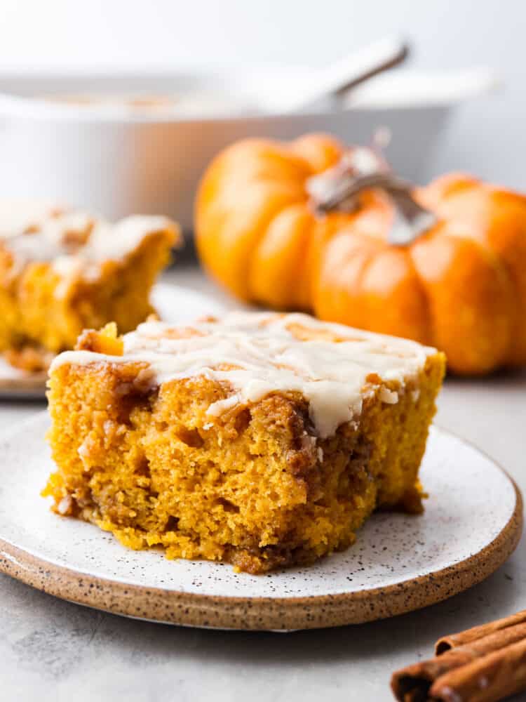 This pumpkin cinnamon roll cake is a perfectly moist pumpkin cake with pockets of swirled cinnamon and brown sugar topped with a maple glaze. It tastes just like you are eating a cinnamon roll! This is one of the best fall cakes! A slice of 0umpkin cinnamon roll cake on a speckled plate with small pumpkins in the background of the picture.