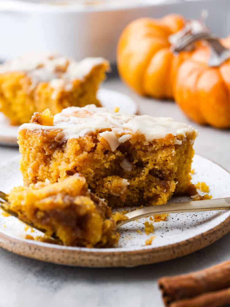 This pumpkin cinnamon roll cake is a perfectly moist pumpkin cake with pockets of swirled cinnamon and brown sugar topped with a maple glaze. It tastes just like you are eating a cinnamon roll! This is one of the best fall cakes! A slice of pumpkin cinnamon roll cake on a plate with a gold fork cutting a bite out of it.