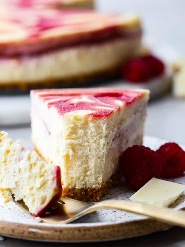 Creamy, rich, and filled with flavor, this white chocolate raspberry cheesecake is a deliciously decadent dessert! Its marble swirl on top and graham cracker crust on the bottom will make you want to serve this dessert with every meal! The side view of a slice of white chocolate raspberry cheesecake on a plate with a fork that cut a bite out.