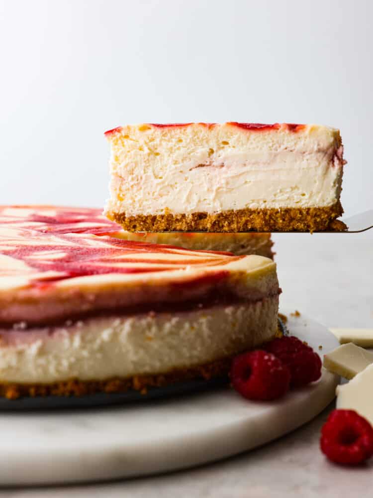 Creamy, rich, and filled with flavor, this white chocolate raspberry cheesecake is a deliciously decadent dessert! Its marble swirl on top and graham cracker crust on the bottom will make you want to serve this dessert with every meal! The side view of a full raspberry cheesecake with a slice being removed.