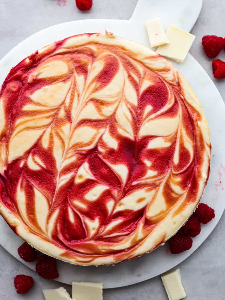 Creamy, rich, and filled with flavor, this white chocolate raspberry cheesecake is a deliciously decadent dessert! Its marble swirl on top and graham cracker crust on the bottom will make you want to serve this dessert with every meal! The top view of a white chocolate raspberry cheesecake on a platter.