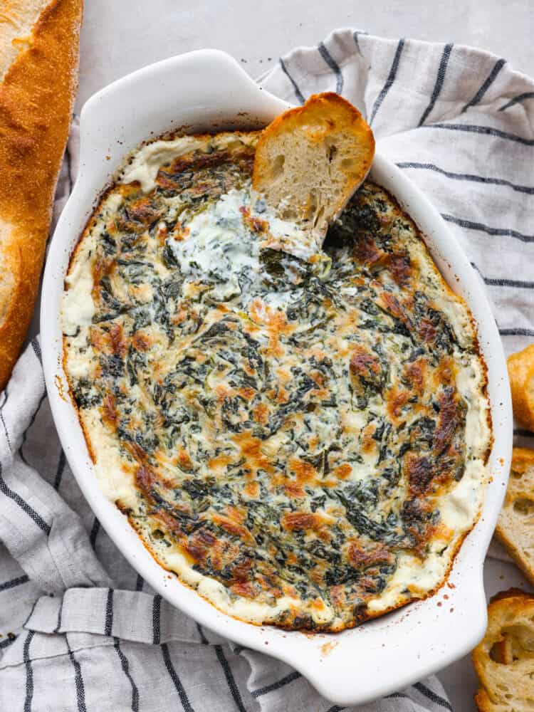 Spinach dip is a classic appetizer known for its creamy and comforting flavors. The combination of spinach, cheeses, and seasonings creates a delicious dip you'll want to bring to all of your gatherings! Top view of spinach dip in a oval white baking dish. A striped dish towel, baguette, and baguette slices are garnished on the side. A baguette slice is dipped in the spinach dip.