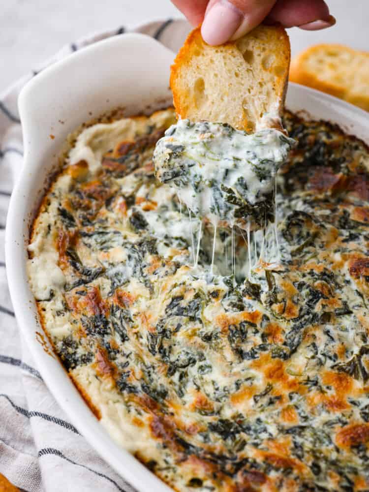 Spinach dip is a classic appetizer known for its creamy and comforting flavors. The combination of spinach, cheeses, and seasonings creates a delicious dip you'll want to bring to all of your gatherings! A close up photo of the baked spinach dip and a baguette slice dipping into the dip showing the cheesy dip lifting out. The baking dish is on top of a striped kitchen towel.
