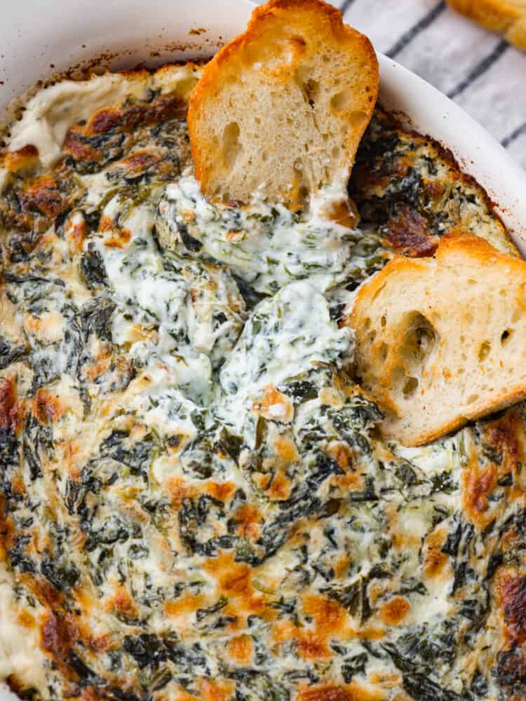 Spinach dip is a classic appetizer known for its creamy and comforting flavors. The combination of spinach, cheeses, and seasonings creates a delicious dip you'll want to bring to all of your gatherings! A close up photo of the baked spinach dip and two baguette slices dipping into the dip.