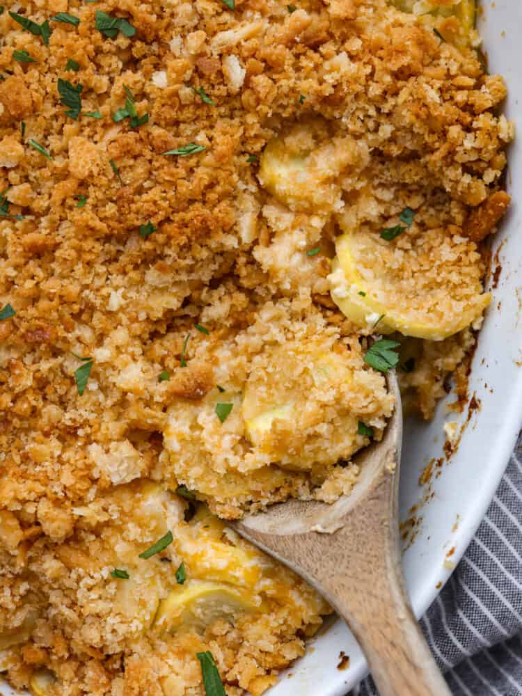 This recipe is the best way to use up that yellow summer squash!ย Squash casserole is a great way to use up fresh squash from your garden. Easy to make with a creamy, cheesy filling and a crispy, buttery topping! A close up of squash casserole with a wooden spoon.