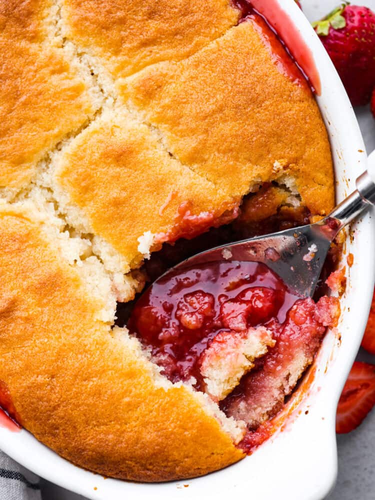 This easy-to-make strawberry cobbler is loaded up with fresh strawberries and topped with a sweet vanilla cake. It's a crisp crust on the outside and is soft and buttery on the inside. Top with some vanilla ice cream for the ultimate treat! Strawberry cobbler in a baking dish with a large spoon scooping some out.