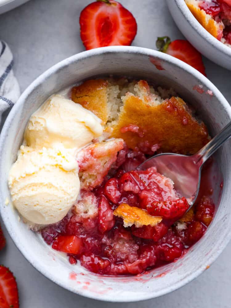 This easy-to-make strawberry cobbler is loaded up with fresh strawberries and topped with a sweet vanilla cake. It's a crisp crust on the outside and is soft and buttery on the inside. Top with some vanilla ice cream for the ultimate treat! The top view of strawberry cobbler in a bowl with a spoon and some vanilla ice cream.