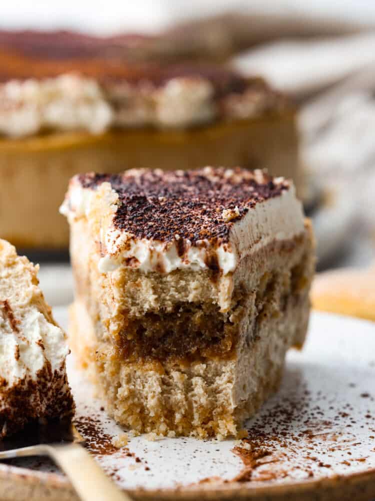 This decadent and delicious tiramisu cheesecake is creamy and will melt in your mouth with every bite. Trust me when I tell you that you will want to make this dessert every time you have guests! A slice of tiramisu cheesecake on a plate with a bite taken out of it.