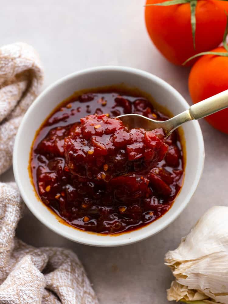 Tomato chutney is a sweet and savory condiment bursting with tomato flavor, delicious spices, vinegar, and brown sugar. Best known for being the perfect pairing for Indian cuisine, tomato chutney is a great addition to curries, naan bread, and charcuterie boards. Top view of tomato chutney in a white bowl with a gold spoon lifting up chutney. A tan kitchen towel and whole tomatoes are next to the bowl of chutney.